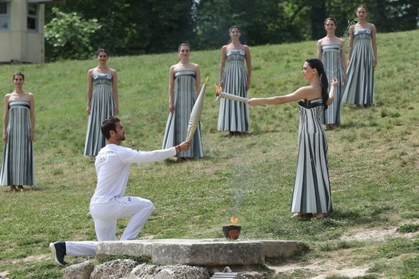 扮演最高女祭司的演員瑪麗·米娜（前右）點燃第一棒火炬手——希臘運動員、東京奧運會男子賽艇項目金牌得主斯特凡諾斯·恩圖斯科斯手中的火炬。新華社記者 趙丁喆 攝